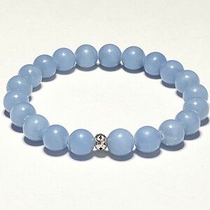 May include: A light blue beaded bracelet. The bracelet features round, smooth beads strung together. A decorative silver bead with a floral design is centered on the bracelet. The beads are a soft, pastel blue color.