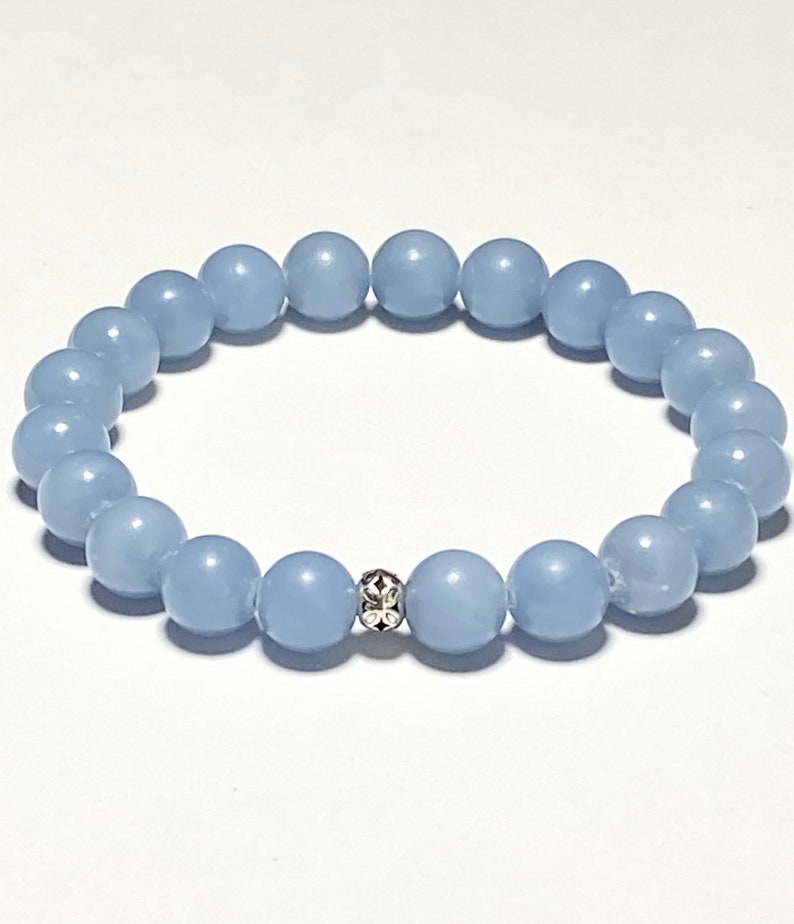 May include: A light blue beaded bracelet. The bracelet features round, smooth beads strung together. A decorative silver bead with a floral design is centered on the bracelet. The beads are a soft, pastel blue color.