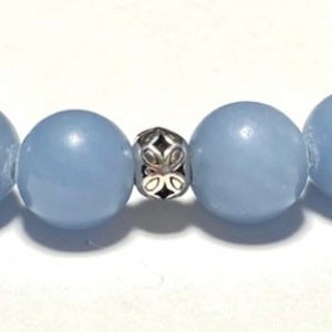 May include: Close-up of a bracelet featuring large, round, pale blue beads. A decorative silver-colored bead with an openwork design is centered between the blue beads. The bracelet is on a white surface.