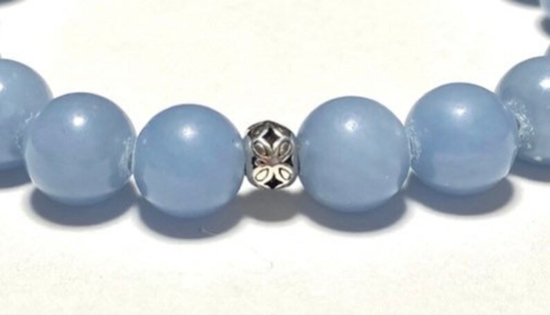 May include: Close-up of a bracelet featuring large, round, pale blue beads. A decorative silver-colored bead with an openwork design is centered between the blue beads. The bracelet is on a white surface.