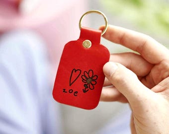 Puede incluir: Un llavero de cuero rojo con un anillo dorado. El llavero tiene un corazón, una flor y el nombre "Zoe" dibujados con tinta negra. El llavero está en la mano de una persona.