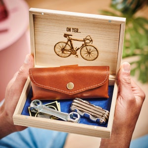 May include: A wooden box containing a leather pouch and a multi-tool. The box lid features a laser-engraved image of a bicycle with the text "On Yer..."