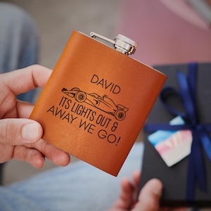 May include: A brown leather flask with a silver cap. The flask is engraved with the name "DAVID", a race car graphic, and the phrase "ITS LIGHTS OUT & AWAY WE GO!"