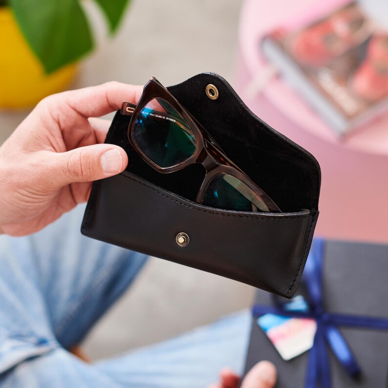 May include: A black leather glasses case with a snap closure, holding a pair of brown-framed eyeglasses. The glasses have a rectangular shape. The case is open, revealing the glasses inside. The background is blurred, with hints of other objects.