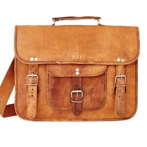May include: A tan leather satchel with a front flap, a handle, and a shoulder strap. The bag features a front pocket with a buckle closure and two additional buckle closures. The initials "M.S.T" are embossed on the front.