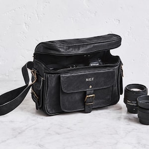 Puede incluir: Una bolsa de cámara de cuero negro con cierre de cremallera superior, correa ajustable y un bolsillo frontal con hebilla de latón y las iniciales M.G.T. La bolsa está abierta, mostrando un cuerpo de cámara en su interior y dos objetivos al lado.