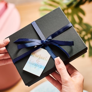 May include: A black gift box tied with a navy blue ribbon and bow. A small tag with the names "Herbert & Winifred" is attached to the ribbon. The box has a textured surface.