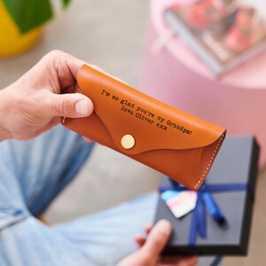 May include: A brown leather glasses case with the text "I'm so glad you're my Grandpa! love Oliver xxx" and a gold button closure. The case is held in front of a gift box with a blue ribbon.