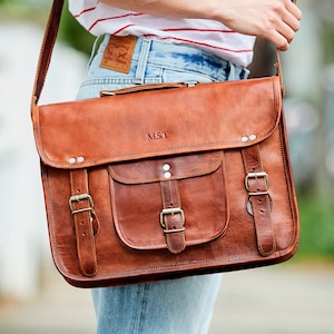 May include: A brown leather messenger bag with a front flap, buckle closures, and a shoulder strap. The bag has a front pocket and the initials "M.S.T." are embossed on the front. The bag is carried over the shoulder.