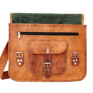 May include: A tan leather satchel with a green interior flap. The bag has a front pocket with a buckle fastening, a zipped top, and a shoulder strap. The initials "M.S.T" are embossed on the front.