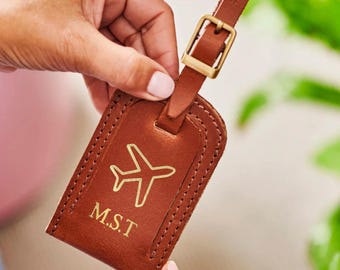 May include: A brown leather luggage tag with a gold airplane graphic and the initials M.S.T. The tag has a stitched border and a matching leather strap with a gold buckle. A travel accessory.