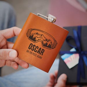 May include: A brown leather flask with a silver cap and hinge. The flask features a black graphic of ski goggles with a mountain scene and the name "OSCAR" with the text "ON THE PISTE AGAIN".