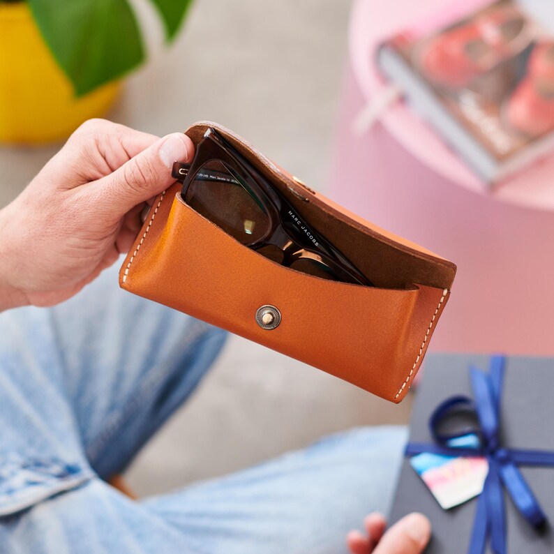 May include: A brown leather glasses case with a pair of eyeglasses inside. The case is open, revealing the glasses. The case has a snap closure and white stitching around the edges. The glasses have black frames. The brand name "MARC JACOBS" is visible on the glasses.