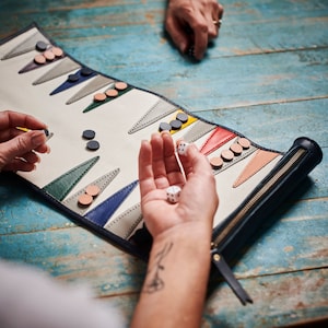 May include: A rolled-up backgammon board with a cream-colored playing surface and colorful triangular points. The board is trimmed in navy blue with a zipper closure. Dice and game pieces are visible, suggesting a game in progress.