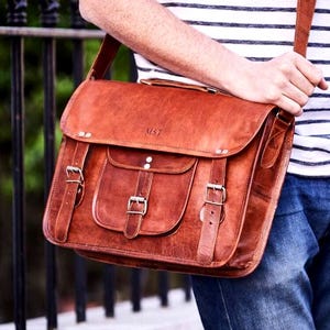 May include: A brown leather messenger bag with a flap closure, two front pockets, and an adjustable shoulder strap. The bag features metal buckles and the initials "MST" embossed on the front. The bag is held by a person wearing a striped top and jeans.
