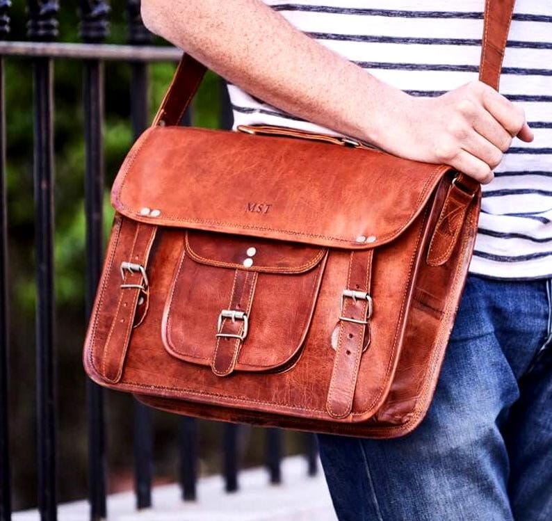 May include: A brown leather messenger bag with a flap closure, two front pockets, and an adjustable shoulder strap. The bag features metal buckles and the initials "MST" embossed on the front. The bag is held by a person wearing a striped top and jeans.
