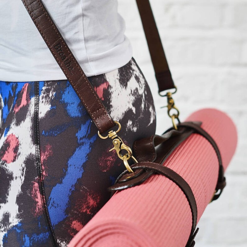 May include: A brown leather yoga mat strap with a brass buckle and the word "Breathe" embossed on the strap. The strap is attached to a rolled up pink yoga mat.