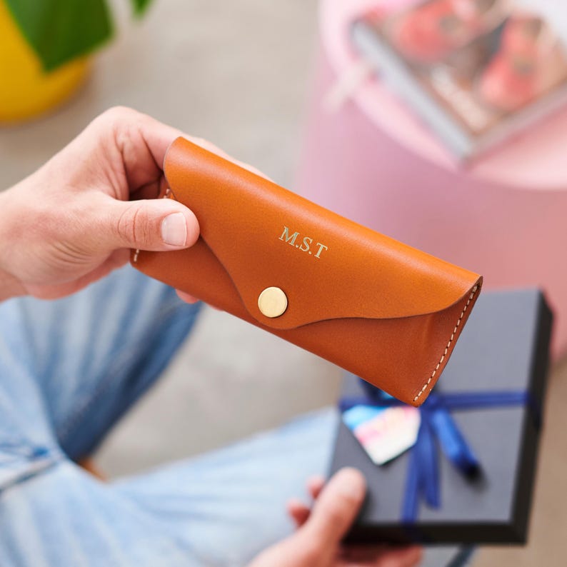 May include: A brown leather glasses case with the initials "M.S.T." embossed in gold. The case has a flap closure with a gold button and white stitching. A gift box with a blue ribbon is in the background.