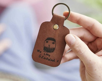 Puede incluir: Un llavero de cuero marrón con un dibujo de una persona con barba y el texto "We Love Granddad X" grabado en él.