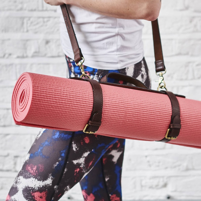 May include: A pink yoga mat with brown leather straps and gold buckles. The mat is rolled up and being carried by a person.