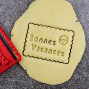 Peut inclure: Emporte-pièce en plastique rouge avec les mots "Bonnes Vacances" et un soleil avec des lunettes de soleil. L'emporte-pièce est posé sur une feuille de pâte à biscuits non cuite avec le même motif découpé.