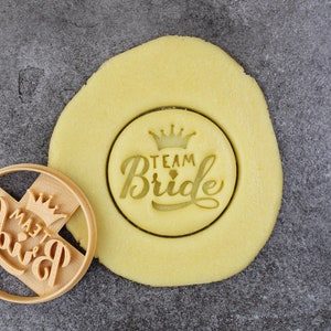 Peut inclure: Un emporte-pièce en bois avec l'inscription "Team Bride" et un motif de couronne. L'emporte-pièce est utilisé pour créer un biscuit avec le même motif. La pâte à biscuits est de couleur jaune clair.