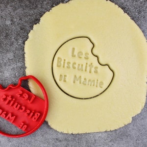 May include: A red plastic cookie cutter in the shape of a bitten cookie with the text "Les Biscuits De Mamie" on it. The cookie cutter is next to a round piece of cookie dough with the same text imprinted on it.