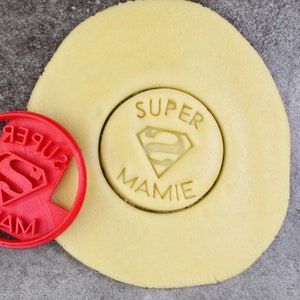 May include: Red plastic cookie cutter with the words "Super Mamie" and a Superman logo. A plain round cookie is next to the cutter.