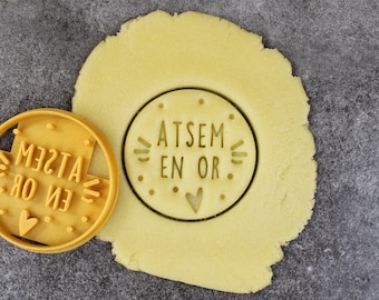 May include: A yellow cookie cutter with the text "ATSEM EN OR" and a small heart. The cookie cutter is on top of a round piece of unbaked cookie dough.