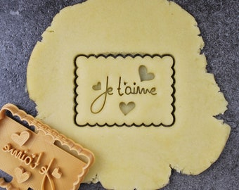 May include: A brown plastic cookie cutter with the words "Je t'aime" and hearts. The cutter is on top of a round piece of unbaked cookie dough.