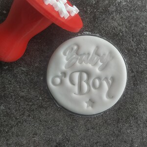 May include: White round cookie cutter with the words "Baby o' Boy" embossed on it. The cutter is on a grey surface next to a red plastic handle.