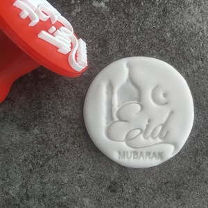 Può includere: Un timbro di plastica bianco con le parole "Eid Mubarak" e un disegno di una moschea. Il timbro viene utilizzato per creare un'impressione su una superficie bianca.
