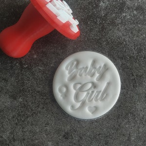 May include: White plastic baby girl cookie stamp with the words "Baby Girl" and a female symbol. The stamp is on a gray surface.