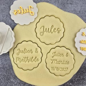 Può includere: Una collezione di timbri per biscotti e un pezzo di pasta. I timbri sono bianchi con bordi smerlati e testi come "Jules", "Julien & Mathilde" e "15/10/2027". Un timbro è una pressa. Il testo è giallo.
