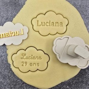 Op de afbeelding: Wolkvormige koekjessnijders en stempels met de naam "Luciana" en het nummer "29" op deeg gedrukt. Eén stempel heeft de naam achterstevoren. De koekjessnijders en stempels zijn beige en het deeg is geel.