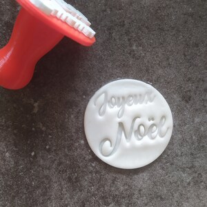 May include: A white plastic cookie stamp with the words "Joyeux Noël" in a cursive font. The stamp is on a grey surface. A red plastic cookie stamp is also visible in the background.