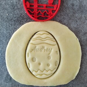 May include: Red plastic cookie cutter in the shape of an Easter egg with the name "Audrey" inside. The cutter is on top of a piece of cookie dough with the cutter's design imprinted into the dough.