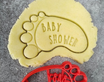 Puede incluir: Cortador de galletas de plástico rojo en forma de pie de bebé con las palabras "BABY SHOWER" en relieve. El cortador está encima de un recorte de masa de galleta amarilla clara.