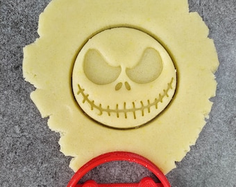 May include: A red plastic cookie cutter shaped like Jack Skellington's face from the Nightmare Before Christmas movie. The cookie cutter is on a gray surface next to a piece of cookie dough with the same shape cut out.