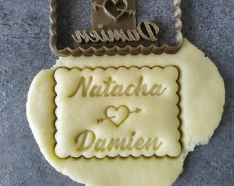 May include: A brown plastic cookie cutter with the words "Natacha" and "Damien" and a heart with an arrow through it. The cookie cutter is on top of a piece of unbaked cookie dough.