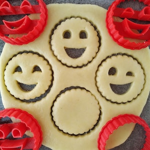 Peut inclure: Des emportes-pièces en plastique rouge en forme de visage souriant, avec un emporte-pièce déjà enfoncé dans une feuille de pâte à biscuits. La pâte est coupée en cinq cercles, dont trois sont coupés en forme de visage souriant.