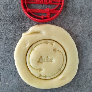 May include: Red plastic cookie cutter with the name Luka and arrows around it. A cookie dough circle with the cutter pressed into it.