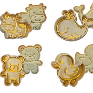 May include: Four cookie cutters shaped like a cow, whale, teddy bear, and duck. Each cutter has a light beige interior and a gold-coloured outline. The cow has yellow spots, and the whale has a water spout.