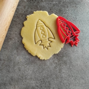 May include: A red plastic rocket cookie cutter on a gray surface with a rolled out piece of cookie dough with the rocket shape cut out.