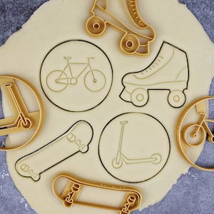 Könnte beinhalten: Ein Satz Ausstecher in Form verschiedener Verkehrsmittel, darunter ein Fahrrad, ein Skateboard, ein Roller, Rollschuhe und ein Tretroller. Die Ausstecher sind aus goldfarbenem Metall und auf einer hellbraunen Oberfläche angeordnet.