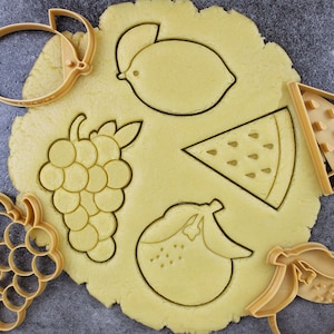Pode incluir: Um close-up de uma massa de biscoito amarela com vários cortadores de biscoitos pressionados na massa. Os cortadores de biscoitos têm a forma de um limão, um cacho de uvas, uma fatia de melancia, uma pêra e um cacho de uvas.