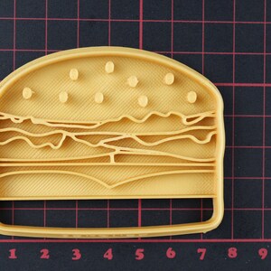 Puede incluir: Un cortador de galletas de pl&aacute;stico amarillo con forma de hamburguesa. El cortador de galletas tiene un dise&ntilde;o detallado con semillas de s&eacute;samo en el pan superior, lechuga, tomate y queso.