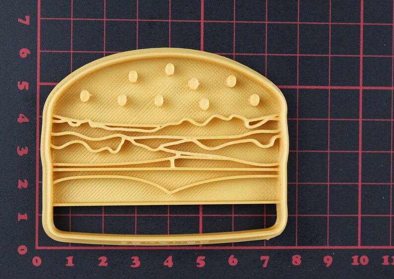 Puede incluir: Un cortador de galletas de pl&aacute;stico amarillo con forma de hamburguesa. El cortador de galletas tiene un dise&ntilde;o detallado con semillas de s&eacute;samo en el pan superior, lechuga, tomate y queso.