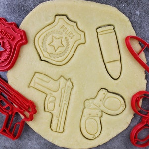Peut inclure: Emporte-pièces en plastique rouge en forme de badge de police, d'un pistolet, de menottes, d'une balle et d'un badge de police avec le mot "POLICE" dessus. Les emporte-pièces sont placés sur une pâte à biscuits ronde.