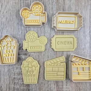 Puede incluir: Un conjunto de cortadores de galletas de plástico beige con formas relacionadas con el cine. Incluyen una cámara de cine, palomitas de maíz, un boleto de cine con la palabra "CINEMA" y una claqueta. Sobre una superficie de madera clara.